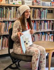 A teen volunteer wearing long pajamas reads a picture book out loud in the Children's Room