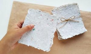 Photo of Caucasian hand holding two sheets of recycled paper (white with colored speckles), with a larger stack wrapped in twine