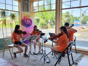 Photo of four teens wearing orange t-shirts, playing string instruments in front of windows and a sign saying "Chamber Music Nor
