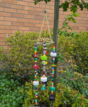 Photo of multi-colored wind chime made from recycled materials and found objects, hanging from tree with bushes and bricks in ba