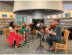 Photo of five musicians in casual clothes performing on stringed instruments in the library.